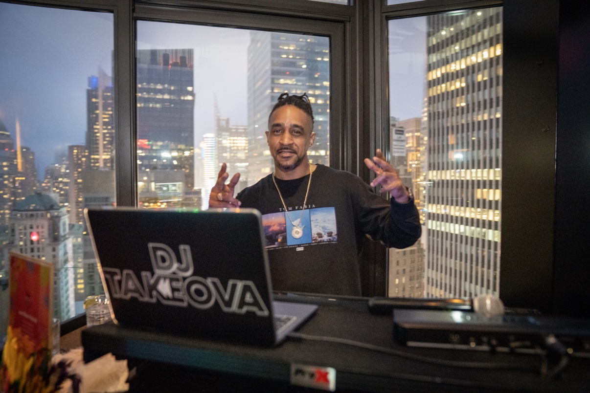 DJ Takeova behind the decks with the NYC skyline at dusk behind him