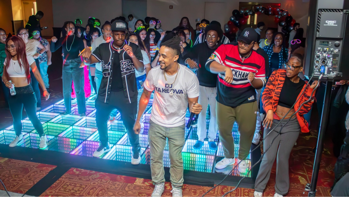 DJ Takeova leading a line dance on a glowing LED dance floor surrounded by students